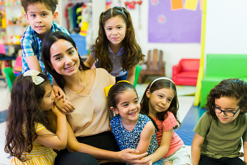 An attractive Latin teacher smiling while hugging and playing with her preschool children students in the classroom